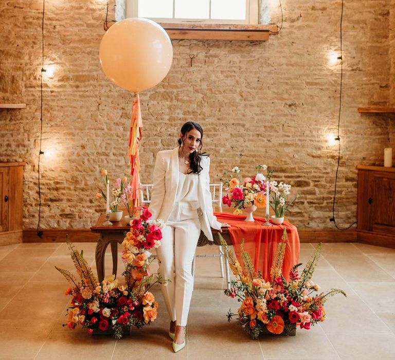 Bride in white bridal suit with closed toe bright green wedding heels standing by metallic pink, white, orange and yellow balloons, wedding fairy lights decorations and wooden table with orange table runner and mixed seasonal flower arrangements 