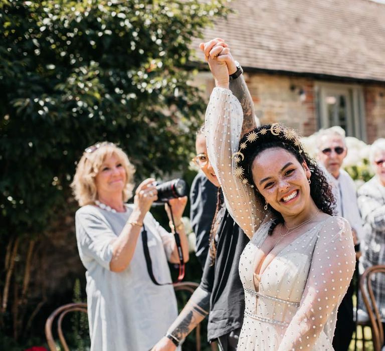 Bride in long sleeve v-neck sheer polka dot overlay sparkly wedding dress and gold celestial bridal headband holding hands with bride smiling into the camera at at-home Viking inspired wedding