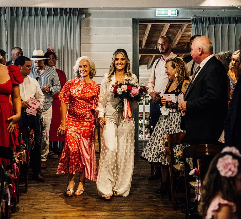 mother of the bride in a pink pattern midi dress walking her daughter down the aisle in a bridal jumpsuit at East Quay wedding 