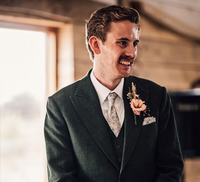 Groom in three piece dark green wedding suit with floral tie and pink flower buttonhole 