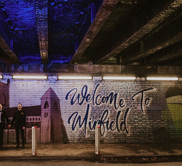 Midfield in Yorkshire wedding with two groom posing next to hand painted mural 