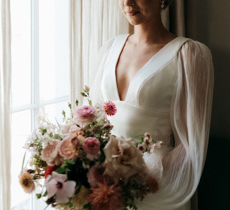 Bride in plunging wedding dress with sheer long sleeves holding pink bouquet 