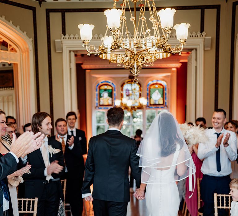 Bride in classic wedding dress from Allure Bridals wearing a wedding veil with trim walking back down the aisle with the groom 