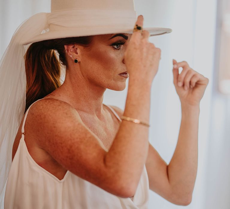 Bride puts on white brimmed wedding hat accessory for boho wedding 