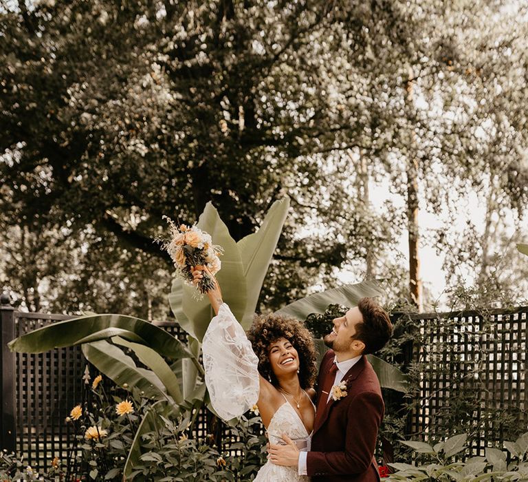 Bride in lace boho wedding dress holding autumnal wedding bouquet with groom in red wedding suit 