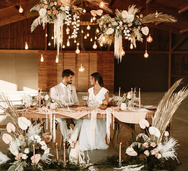 Bride and groom sitting at an intimate tablescape filled with dried flowers, taper candles, natural linens and coloured ceramics under a festoon light installation