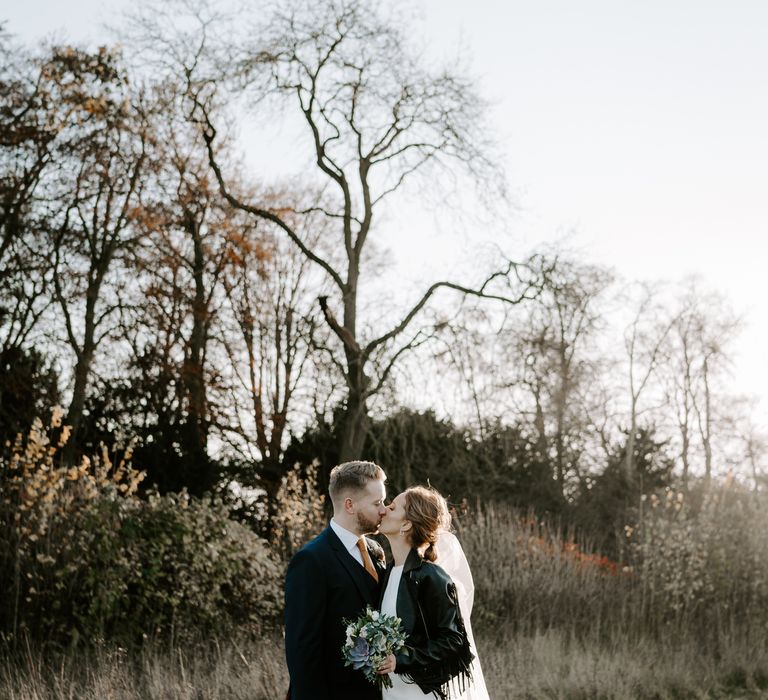 Portrait of the bride and groom kissing with the bride in a fringed leather jacket holding a succulent wedding bouquet 