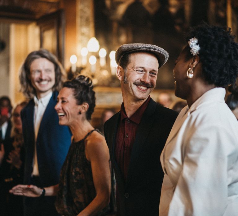 Groom in a red shirt, black suit and flat cap smiling at his bride during the town hall wedding 