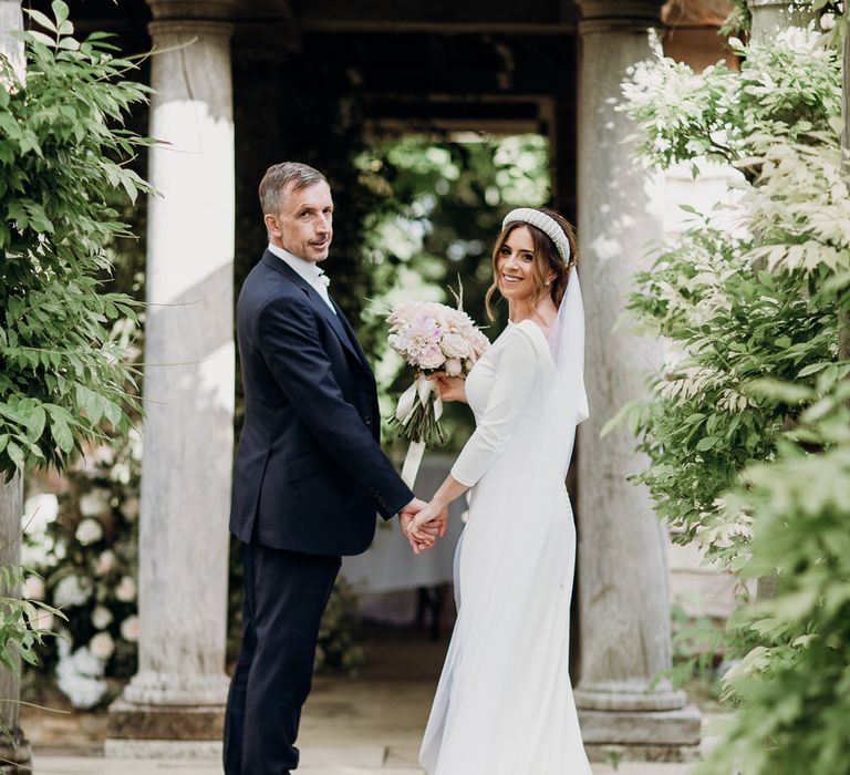 Smiling bride in Elbeth Gillis gown holds blush pink rose bouquet and hand of groom in navy suit in the grounds of Euridge Manor