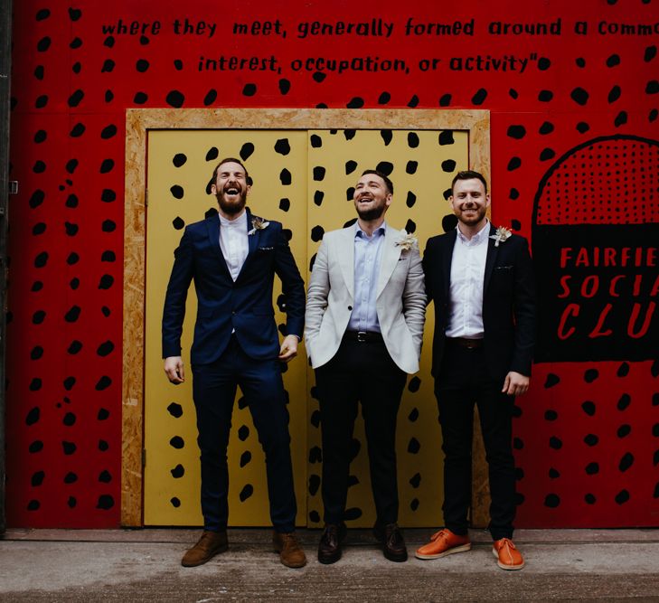 Groom and groomsmen stand laughing in front of red and yellow wall