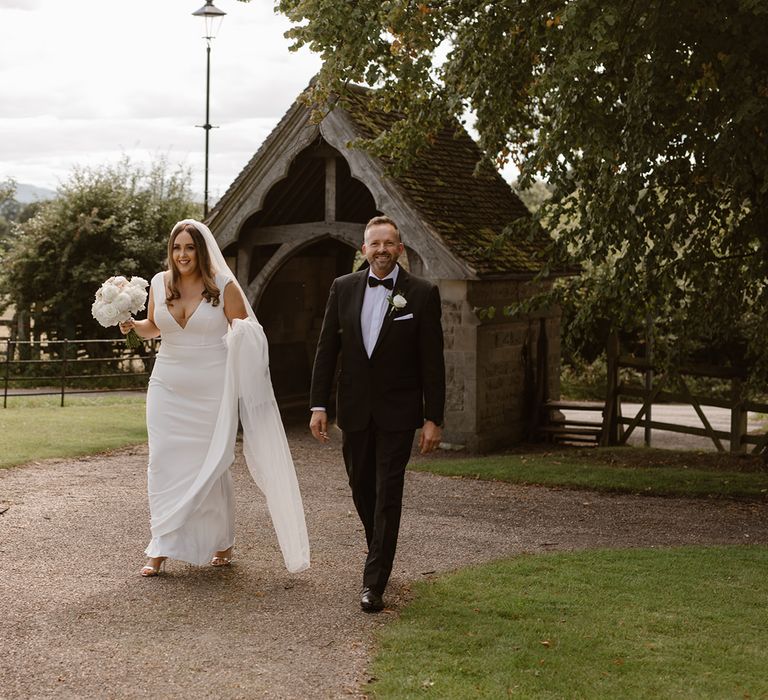 Bride in white Made With Love wedding dress and white chapel length veil holding white and blush pink rose bouquet whilst walking through churchyard with father in black tuxedo