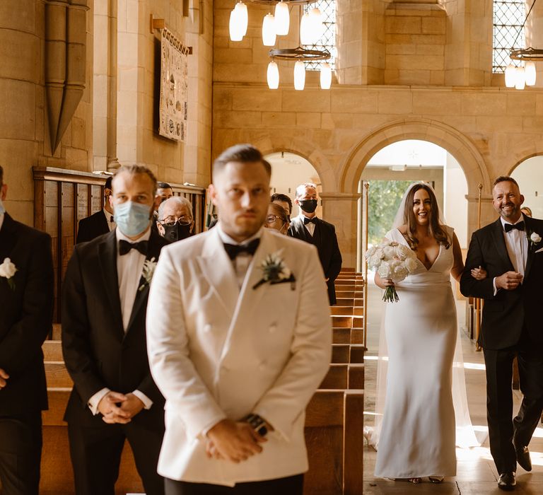 Smiling bride in Made With Love wedding dress walks arm in arm with father in black tuxedo up the church aisle towards groom in white double breasted tuxedo jacket