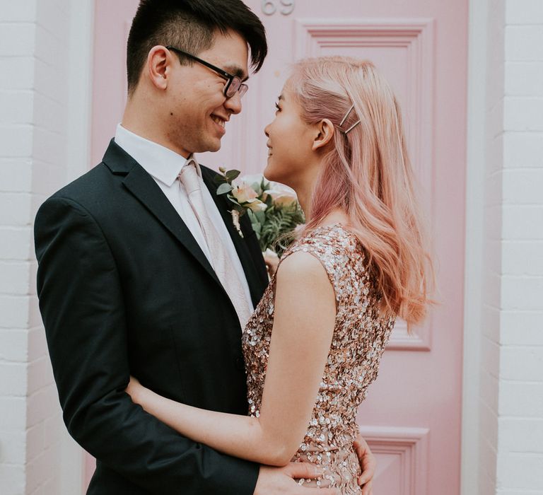 Bride in a pink sequin wedding dress with pink hair standing in front o a pink door with her groom 