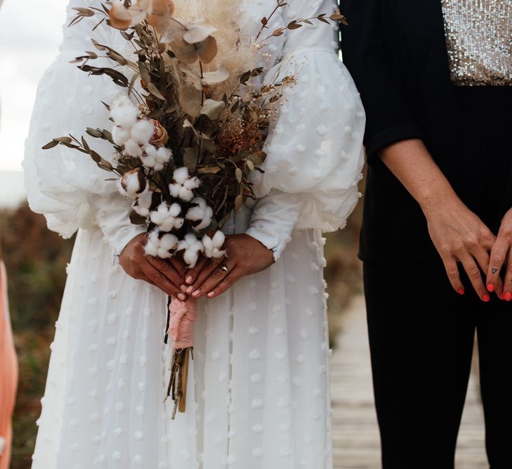 Couple stand side by side, bride in white dress with dried bouquet and bride in black suit and sequin top