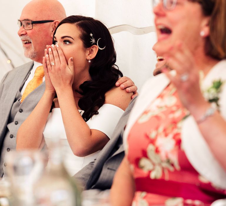 Shocked wedding guests sit with bride wearing celestial hair pins and curled hair holding her hands over her face in shock 