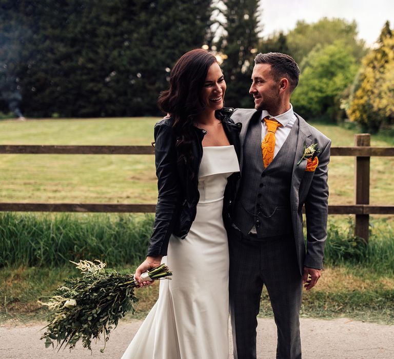 Bride in white Vagabond wedding dress with train and bridal leather jacket hugs groom in grey Hugo Boss suit with orange pocket square and brown shoes outside