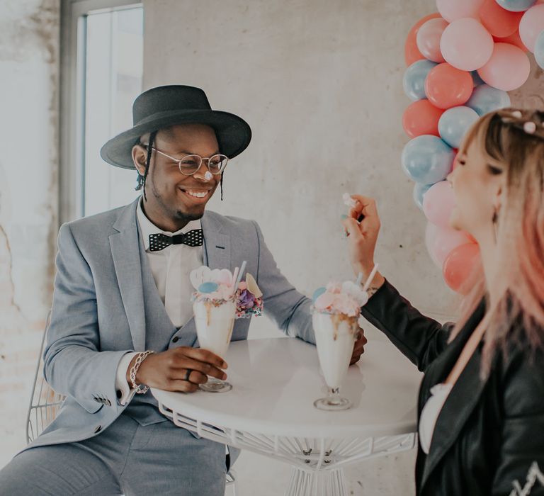 Bride and groom sitting at a table enjoying a milkshake with a Heart balloon installation on the wall 