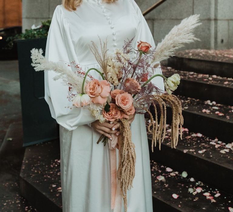 Bride smiles whilst wearing 70s inspired gown and holding floral bouquet with pampas grass