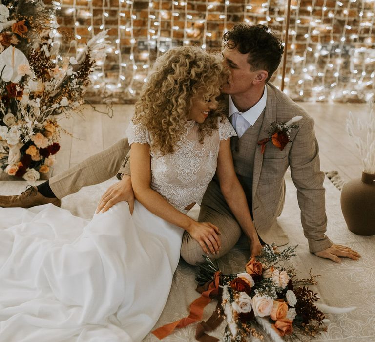 Bride and groom on a beige boho style rug on the floor, in front of a waterfall of fairy lights, surrounded by neutral coloured florals