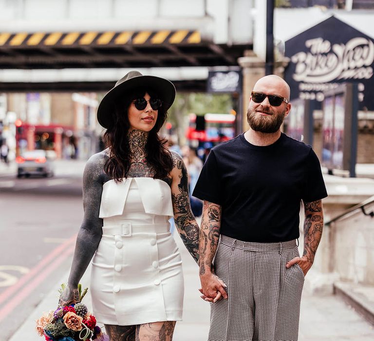 Bride in an alternative wedding dress, bridal boots and fedora hat holding hands with her groom in a check suit and black t-shirt 