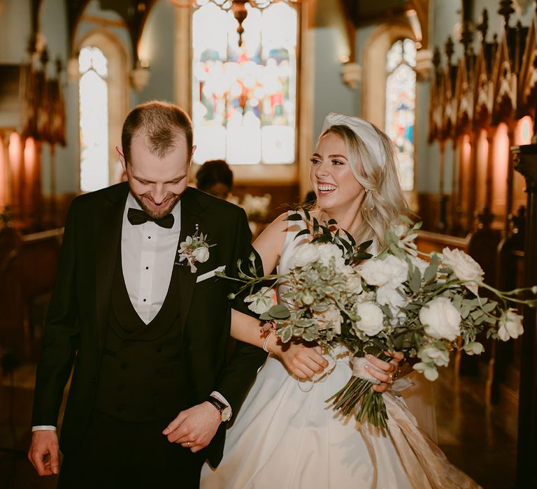 Bride & groom walk down the aisle as bride holds white floral bouquet with green foliage 