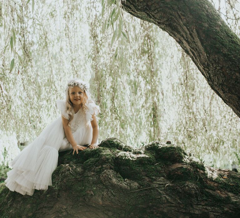 A flower girl in a long white dress and flower crown sits atop a rock at Crab & Lobster fairytale wedding. 