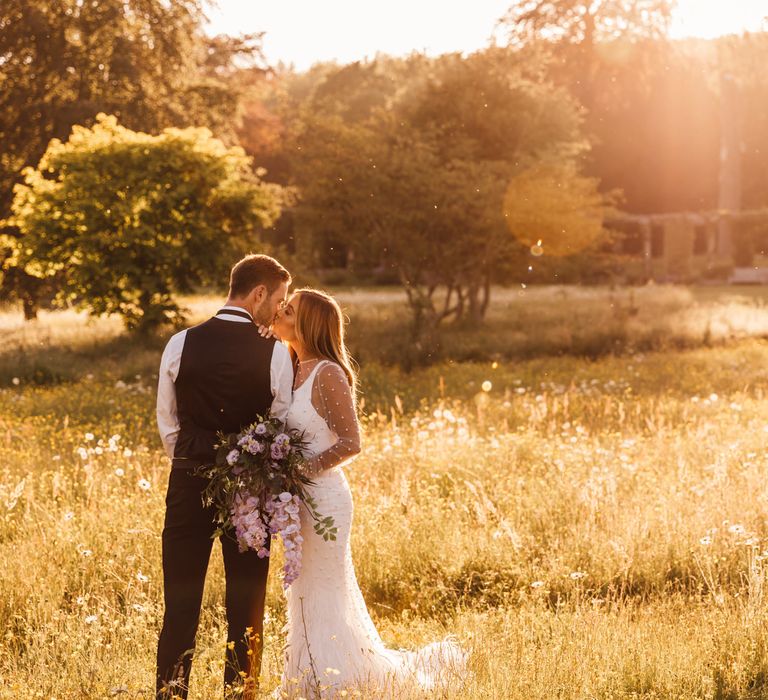 Bride & groom stand within golden fields as the sun sets behind them