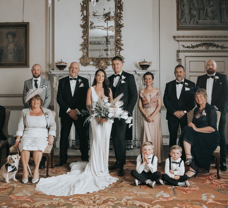 Bride in white Pronovias wedding dress holding white, pink and green wedding bouquet stands with groom in black suit and other wedding guests in formal room at Came House Dorset