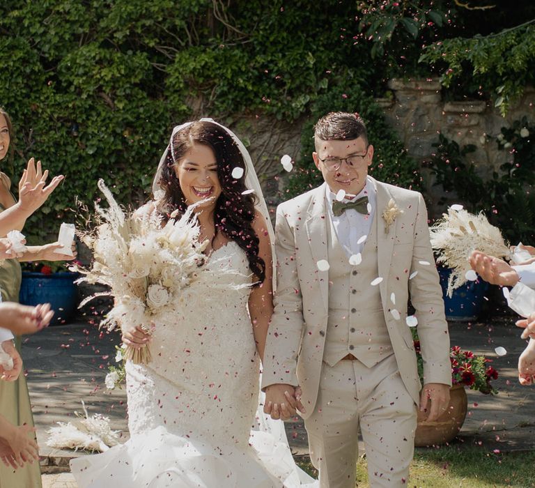 Confetti moment with groom in a beige suit holding hands with his bride in a Mori Lee fishtail wedding dress