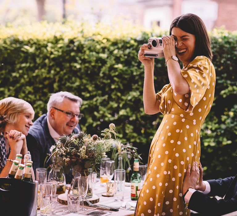 Wedding guest mustard and white polka dot dress taking polaroid pictures during the pub wedding reception 