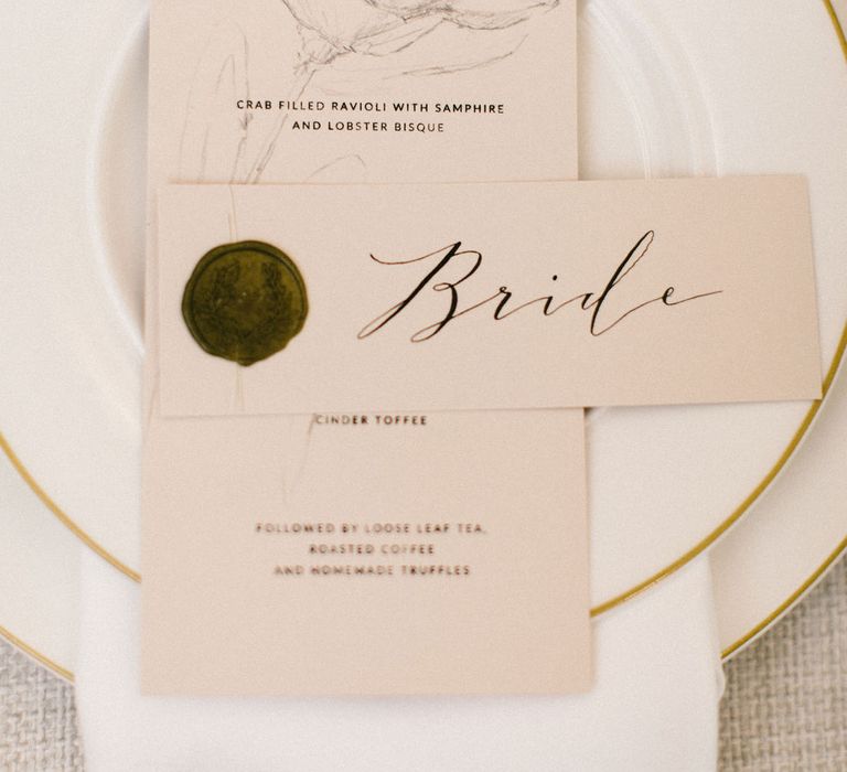 Place setting with gold rimmed white tableware and menu and name place card with wax seal and pink ribbon detail