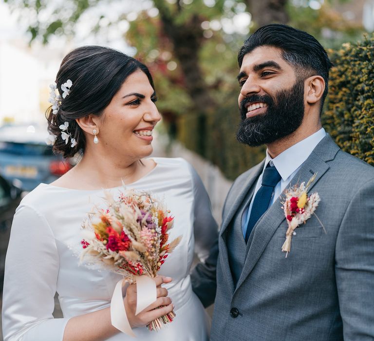 Bride & groom smile as they pose for post-wedding pictures and bride holds vibrant small dried flower bouquet and groom wears matching buttonhole