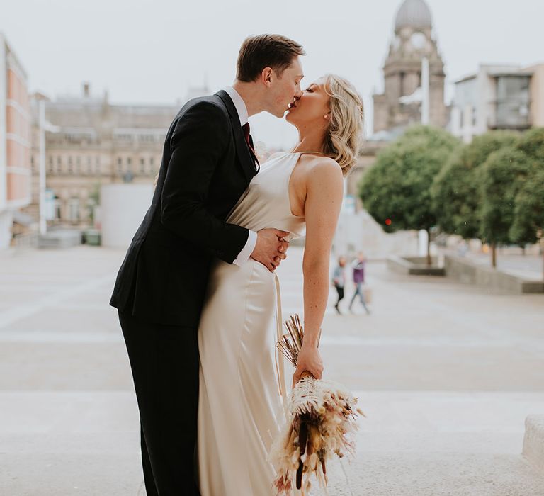 Portrait of the groom in a black suit leaning his bride over for a kiss 