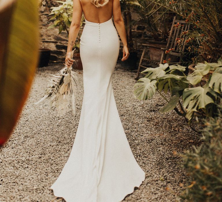 Bride in white open back Made With Love wedding dress with button detailing holding white and green dried bridal bouquet stands in glasshouse at Anran Devon