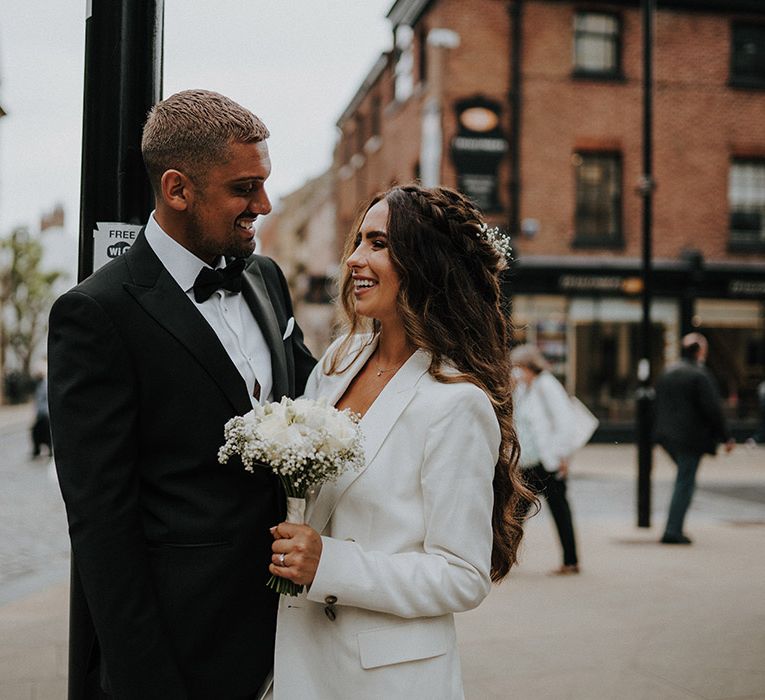 Bride in a white three-piece suit holding a white flower bouquet by a lamppost with her groom in a tuxedo 