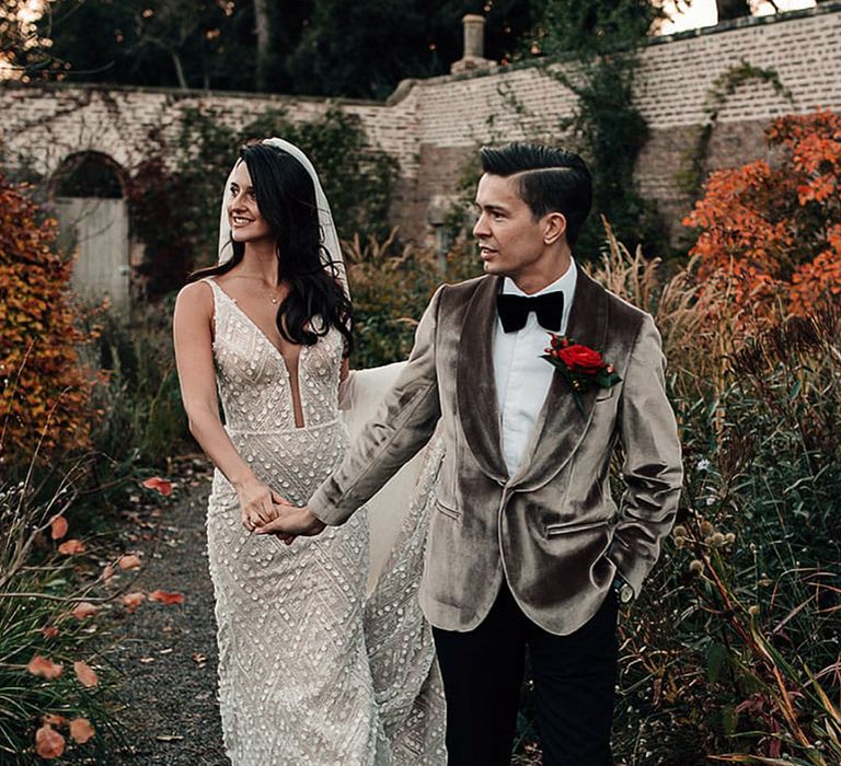 Groom in a grey velvet dinner jacket leading his bride through the gardens at their winter wedding