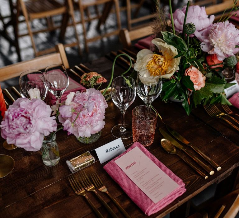 Wedding table place setting on wooden table with pink, yellow and green florals, gold cutlery, stationery by the Noteur and pink napkin for wedding breakfast at Loft Studios London