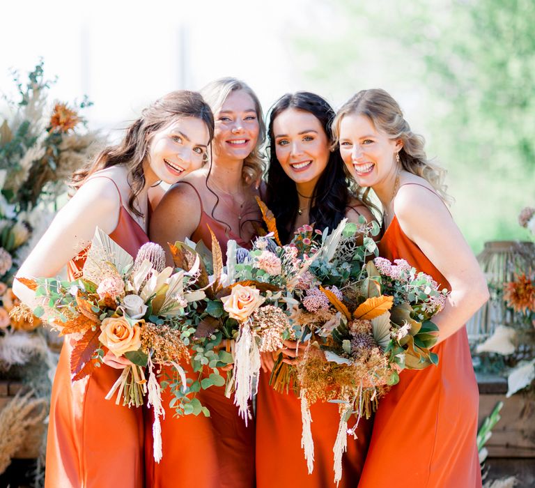 Bridal party portrait with bridesmaid in orange satin bridesmaid dresses holding dried flower bouquets 