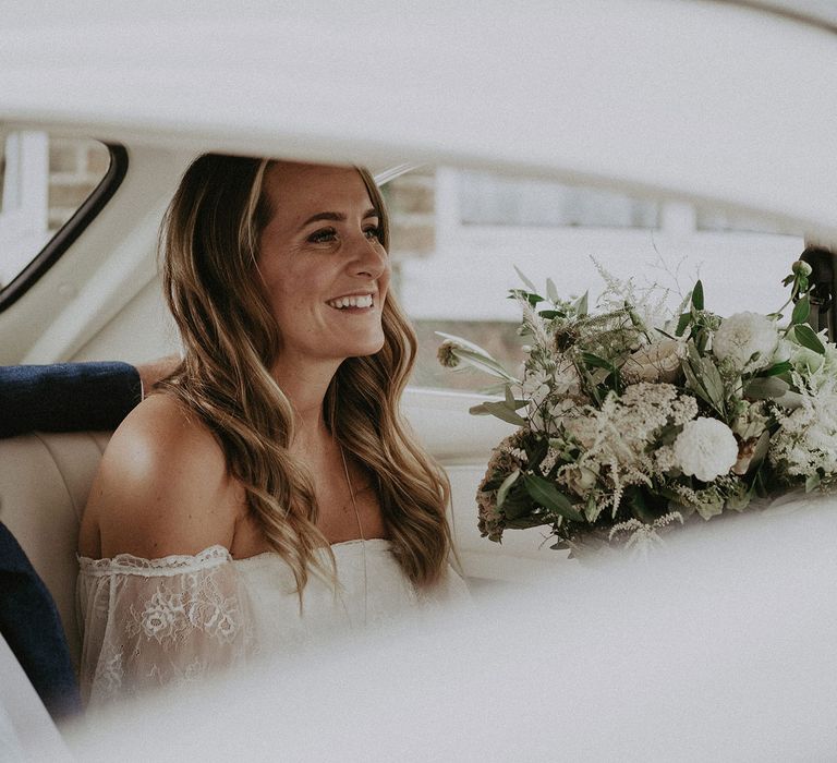 Bride with curled hair wearing Grace Loves Lace bardot wedding dress holding white and green bridal bouquet sits in back of VW bug before Isle of Wight wedding with macrame wedding decor