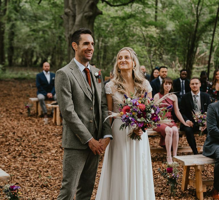 Bride in lace cap sleeved wedding dress holding mixed bridal bouquet stands with groom in grey three piece herringbone suit during woodland wedding ceremony in Norfolk