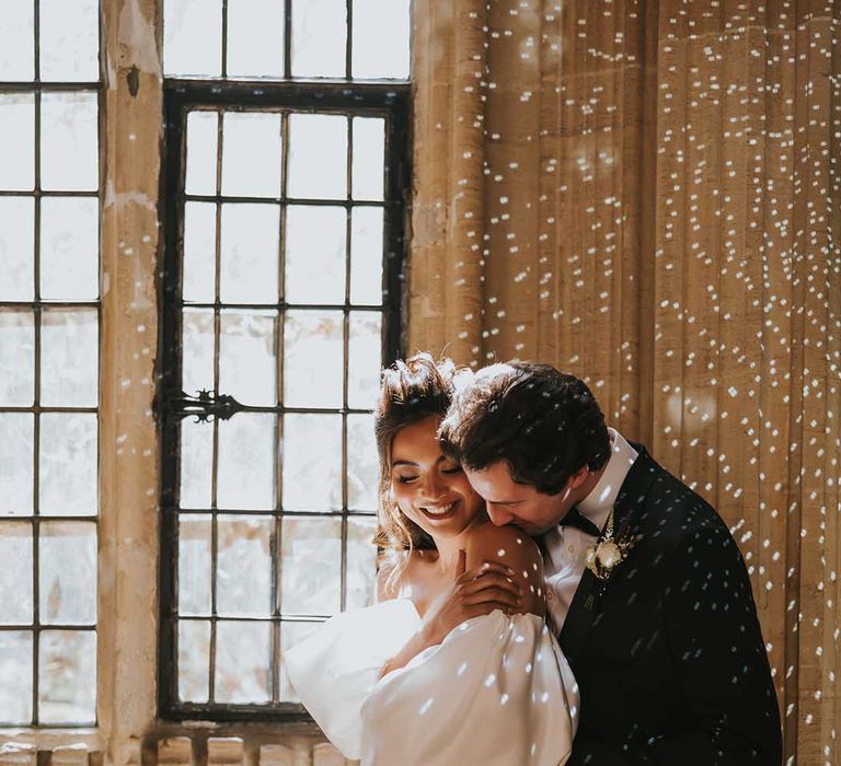 Groom in a tuxedo kissing his brides shoulder in a strapless wedding dress with glistening lights all around them 