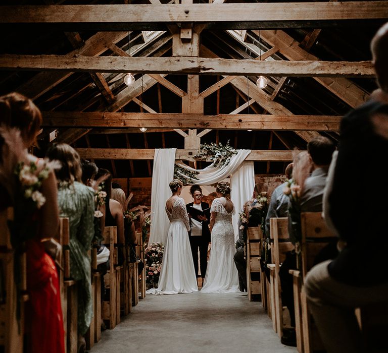 Same-sex wedding ceremony at Woolas Barn with two brides standing at the altar decorated with a drape over the exposed beams 