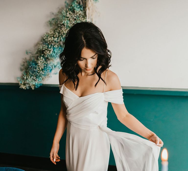 Bride in a cold shoulder wedding dress standing next to plush teal velvet reception chairs 