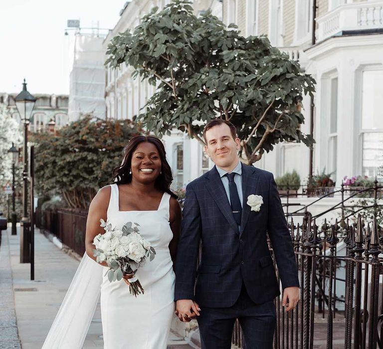 Bride & groom walk together through Chelsea as bride holds white floral bouquet 