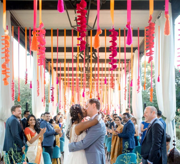 Bride & groom embrace during colourful wedding ceremony surrounded by floral hangings and garlands as wedding guests watch