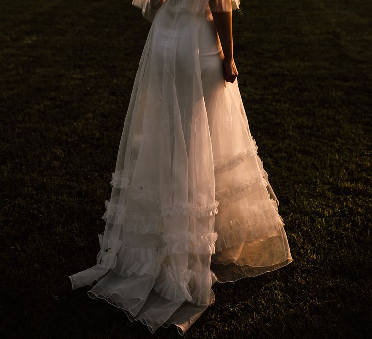 Bride in white Halfpenny London Mayfair dress with riddle sleeves stands in garden during golden hour at Cornish garden wedding reception 