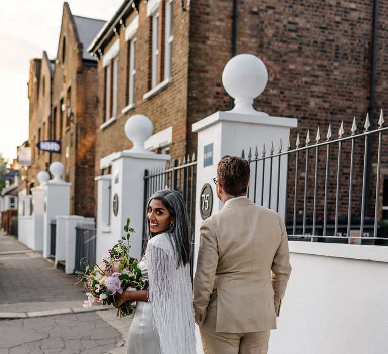 Bride in a feather cape waking through London with her groom in beige suit and gold trainers 