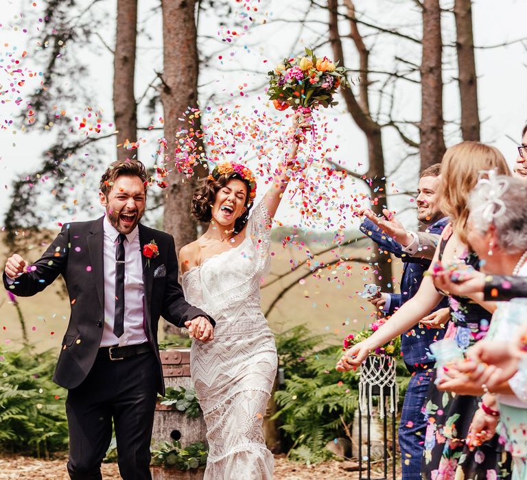 Bride in off the shoulder Grace Loves Lace wedding dress and bridal flower crown holds colourful wedding bouquet and the hand of groom in black suit as guests throw multicoloured confetti