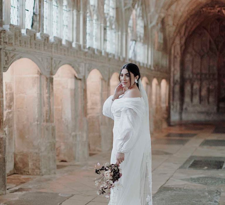 Bride with sleek back hair in an off the shoulder wedding dress with long sleeves and a cathedral length veil with appliqué edge 