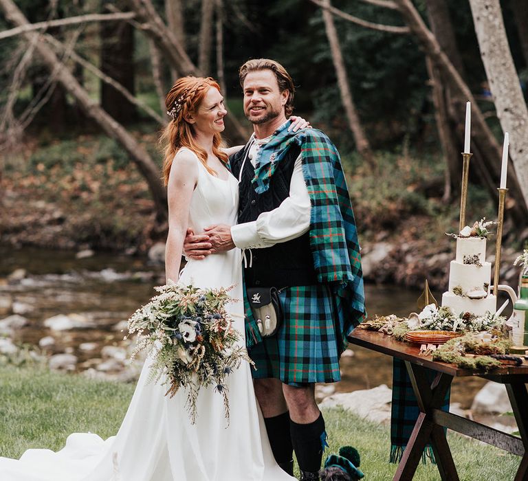 Groom in a kilt and bride in an a-line wedding dress standing next their dessert table with their Scotty dogs by their feet